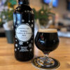 A snifter of dark beer sits atop a Grains & Taps branded coaster. Next to it is a 750 ml amber beer bottle with the main text: Grains & Taps Holiday Cookies & Cream 2024. In the background pine tree holiday decorations are partially in view.