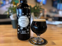 A snifter of dark beer sits atop a Grains & Taps branded coaster. Next to it is a 750 ml amber beer bottle with the main text: Grains & Taps Holiday Cookies & Cream 2024. In the background pine tree holiday decorations are partially in view.