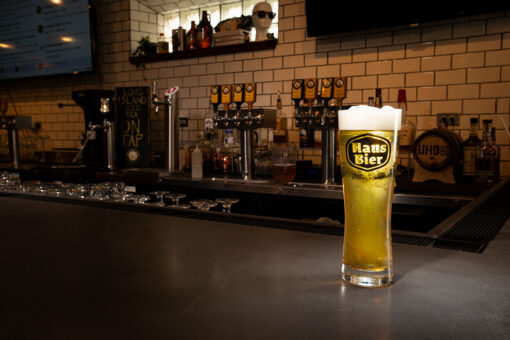 A tall pilsner glass with the lettering "Haus Bier' is prominently positioned on a gray bar top. The beer in the glass is straw colored with white foam at the top. A beam of light is striking the glass. In the dark background are beer taps and various bottles and barwares.