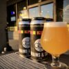 In the foreground a 12 oz tulip glass sits on a gray picnic table. The liquid in the glass is golden with a head of white effervescent carbonation. Behind the glass on the same table is a four pack of 16 oz cans.