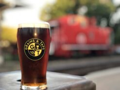 In the foreground, in focus, sunlight hits a pint glass containing a dark red ale. The pint glass is adorned with a round yellow and black Grains & Taps logo. In the background, out of focus is a red train caboose.