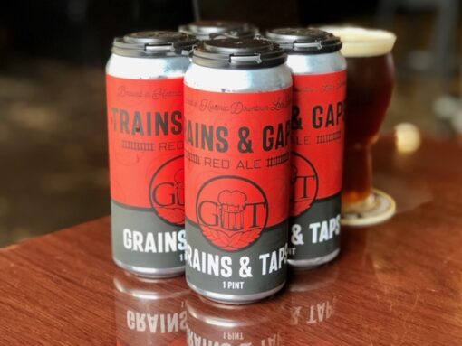 A four pack of 16 oz cans of Trains & Gaps Red Ale from Grains & Taps rests atop a wood table. The labels on the cans have a red base and contain the brewery and beer names. Behind the four pack is a pint glass containing a dark red beer.