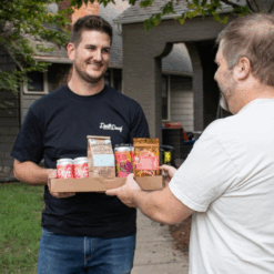 image of a a box of different drinks including coffee beans, beer, and hemp beverages. The box is being handed to the customer in his front yard